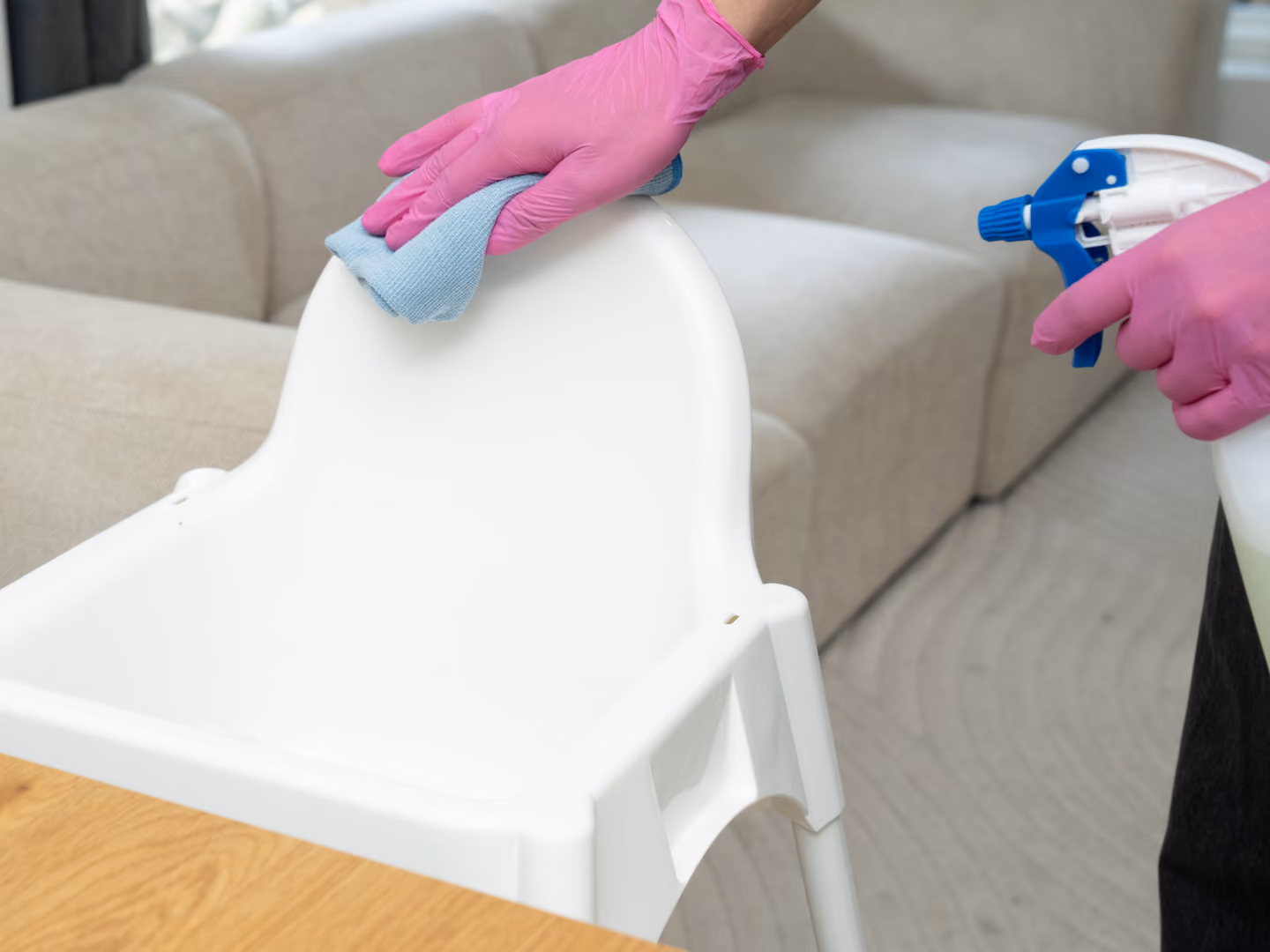 Person wearing pink gloves cleaning a white high chair with a blue cloth and spray bottle.