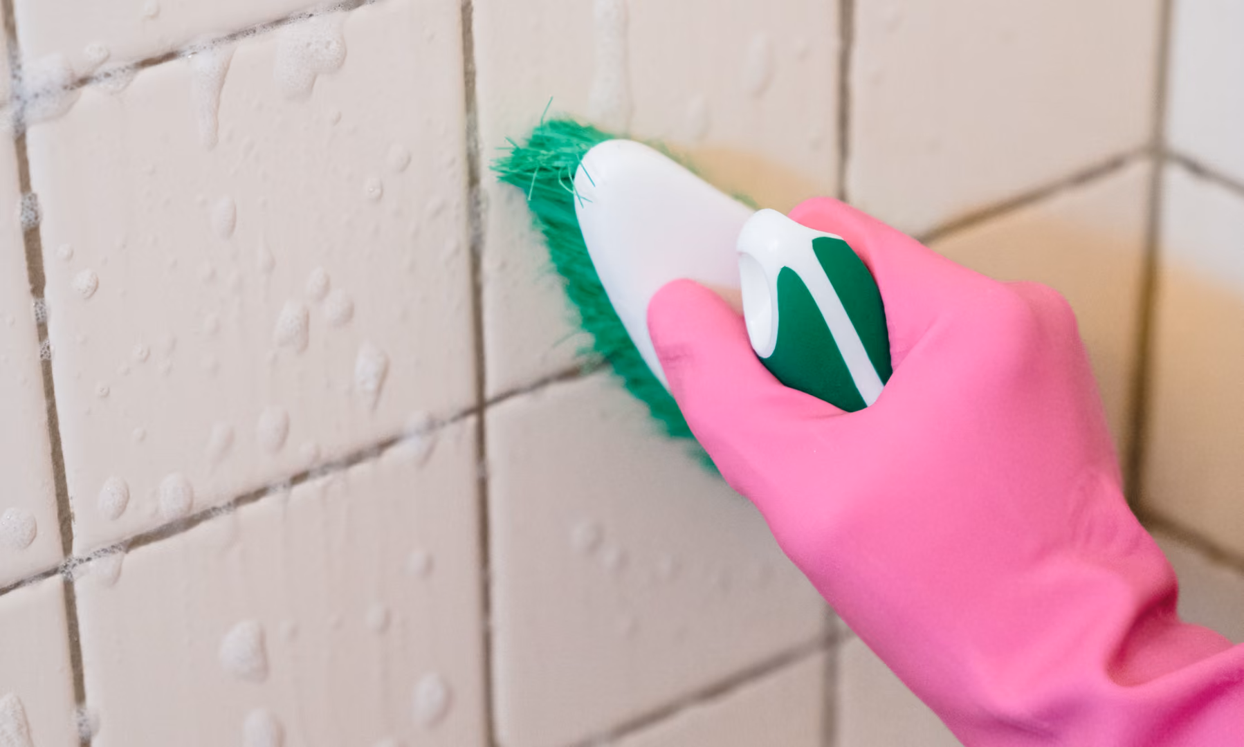 Hand wearing pink glove scrubbing tiled wall grout with green brush and soap suds.