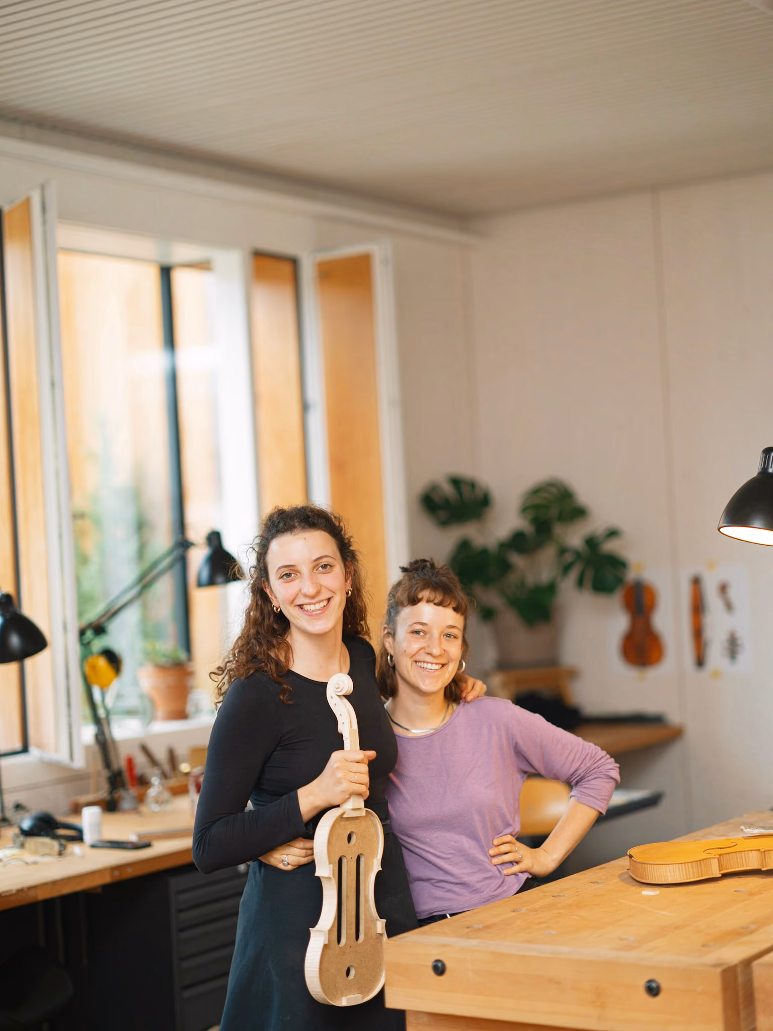 Zwei junge Frauen lächeln, eine hält eine teilweise gefertigte Violine in einer Werkstatt.