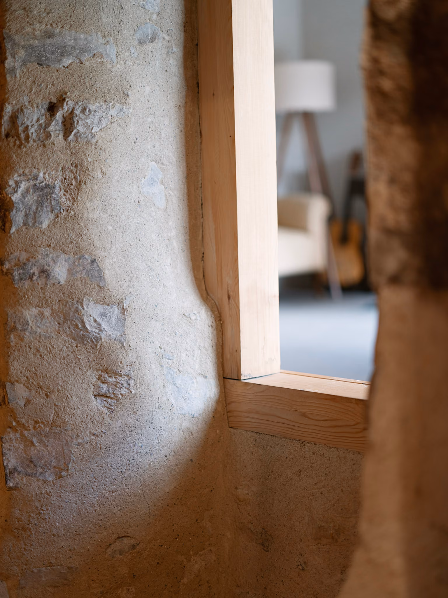Fensterrahmen aus hellem Holz in einer rauen Steinwand mit unscharfen Möbeln im Hintergrund.