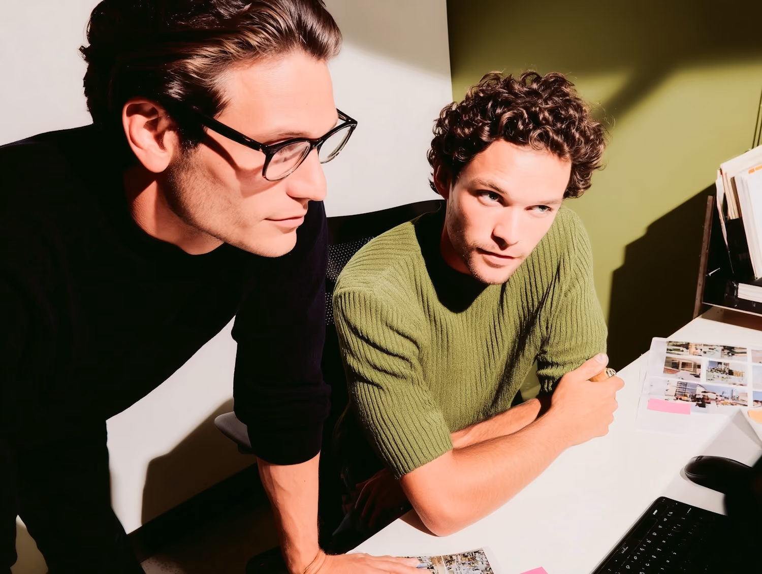 Two men focused on a computer screen, one wearing glasses and the other in a green sweater.
