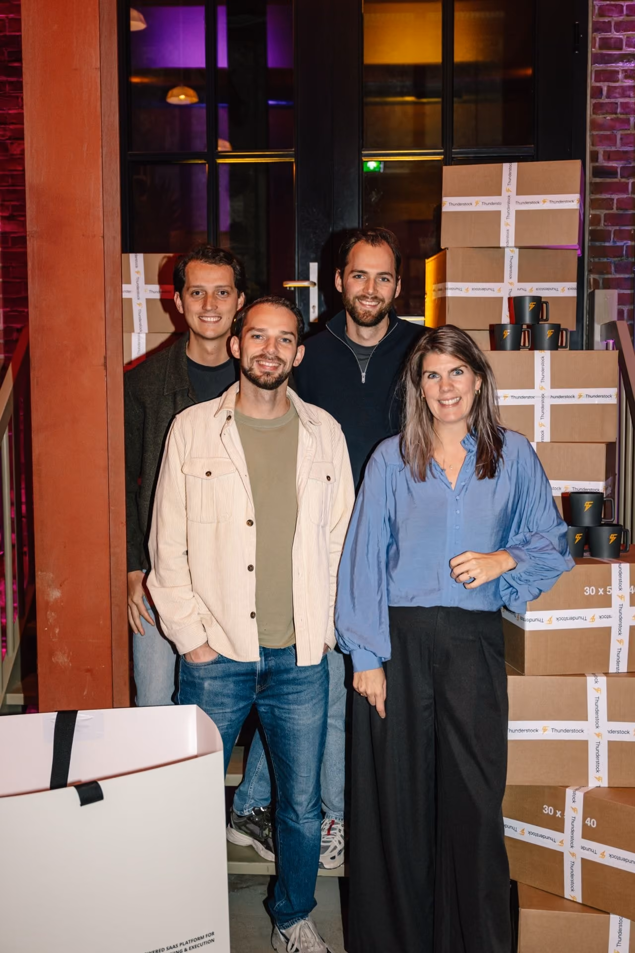 Four smiling adults standing indoors near stacked boxes with Thunderstock logos and branded mugs.