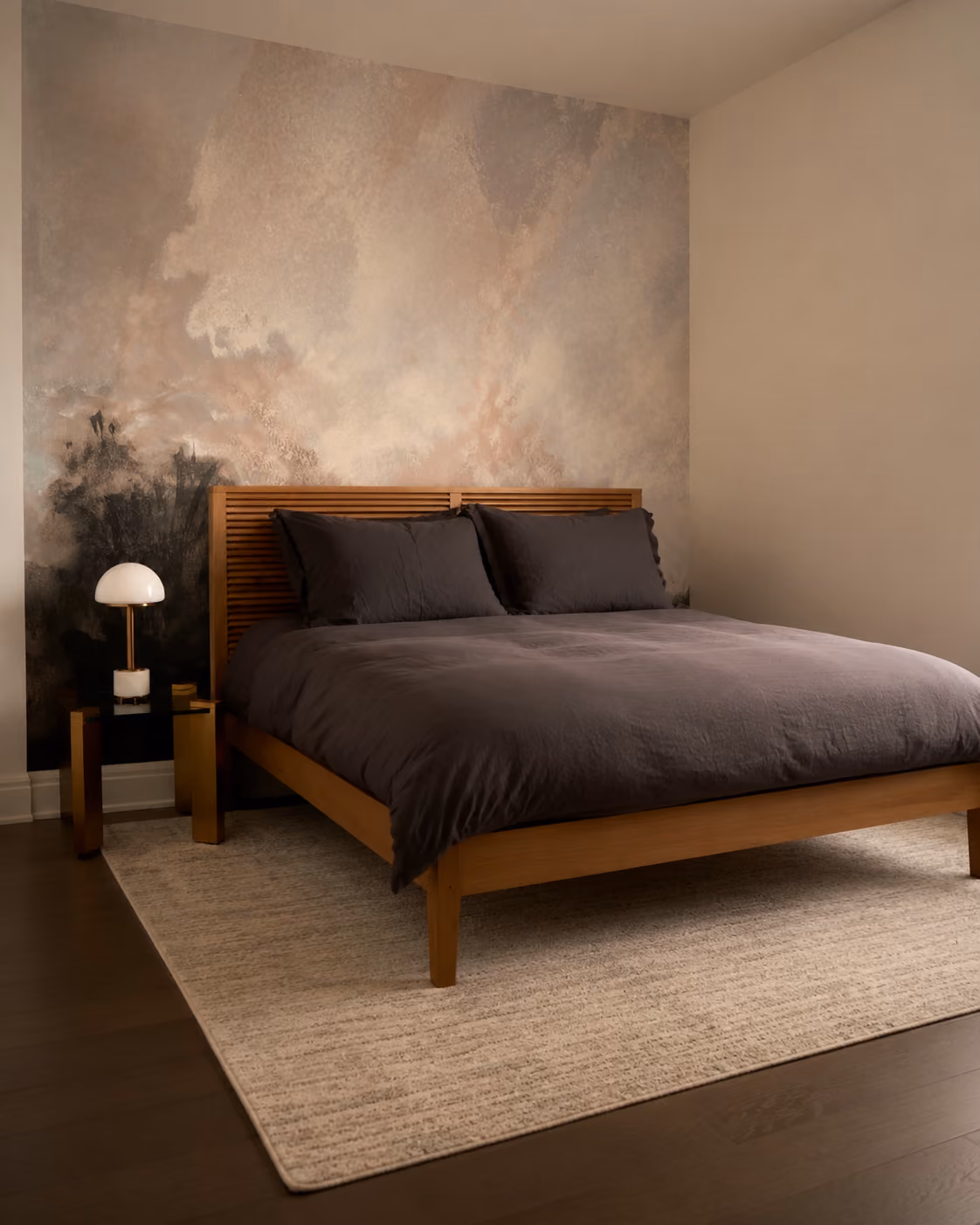 Minimalist bedroom with a wooden bed frame, dark bedding, abstract wall art, and a small lamp on a gold side table.