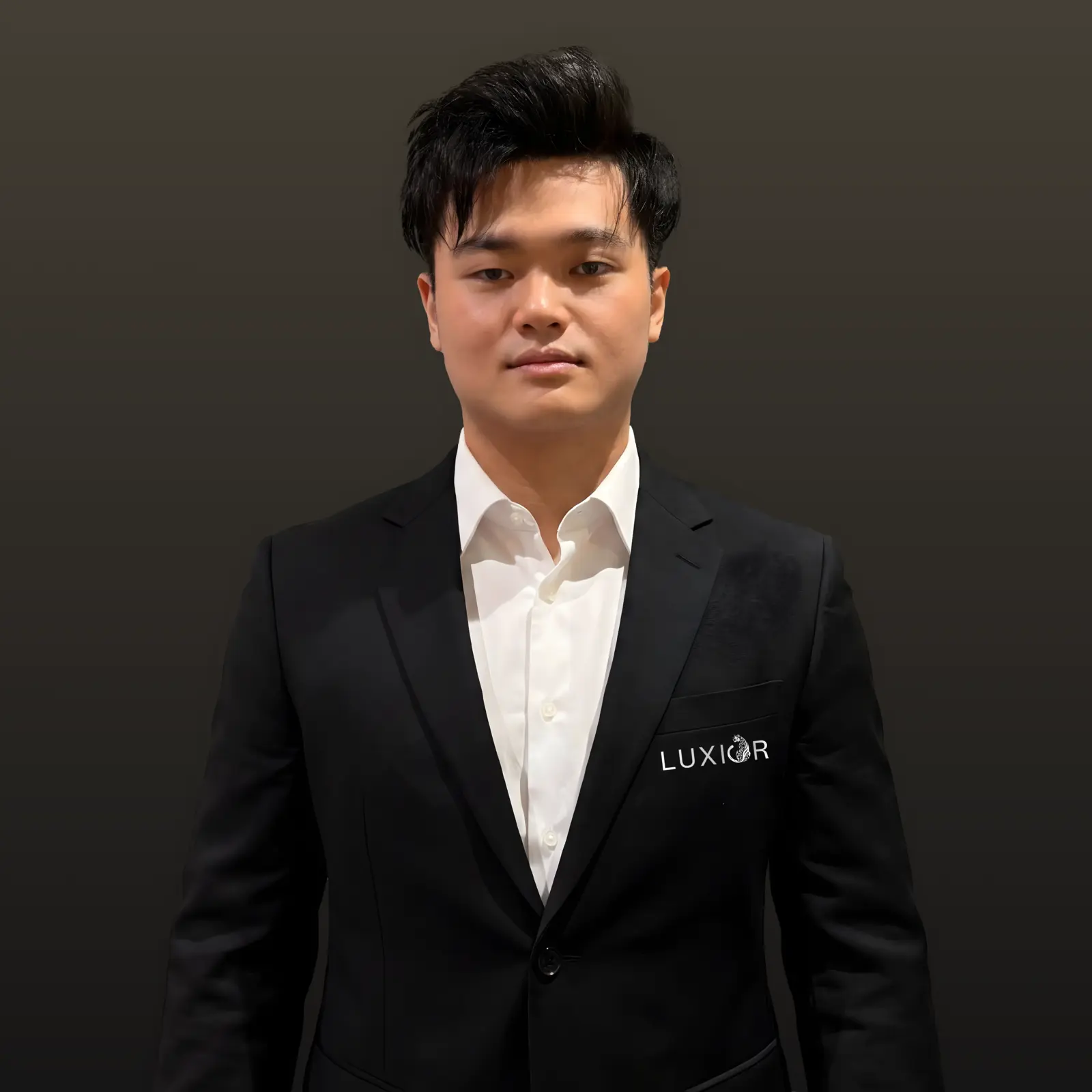 Man with black hair wearing a black suit jacket with LUXIOR logo over a white shirt on dark background.