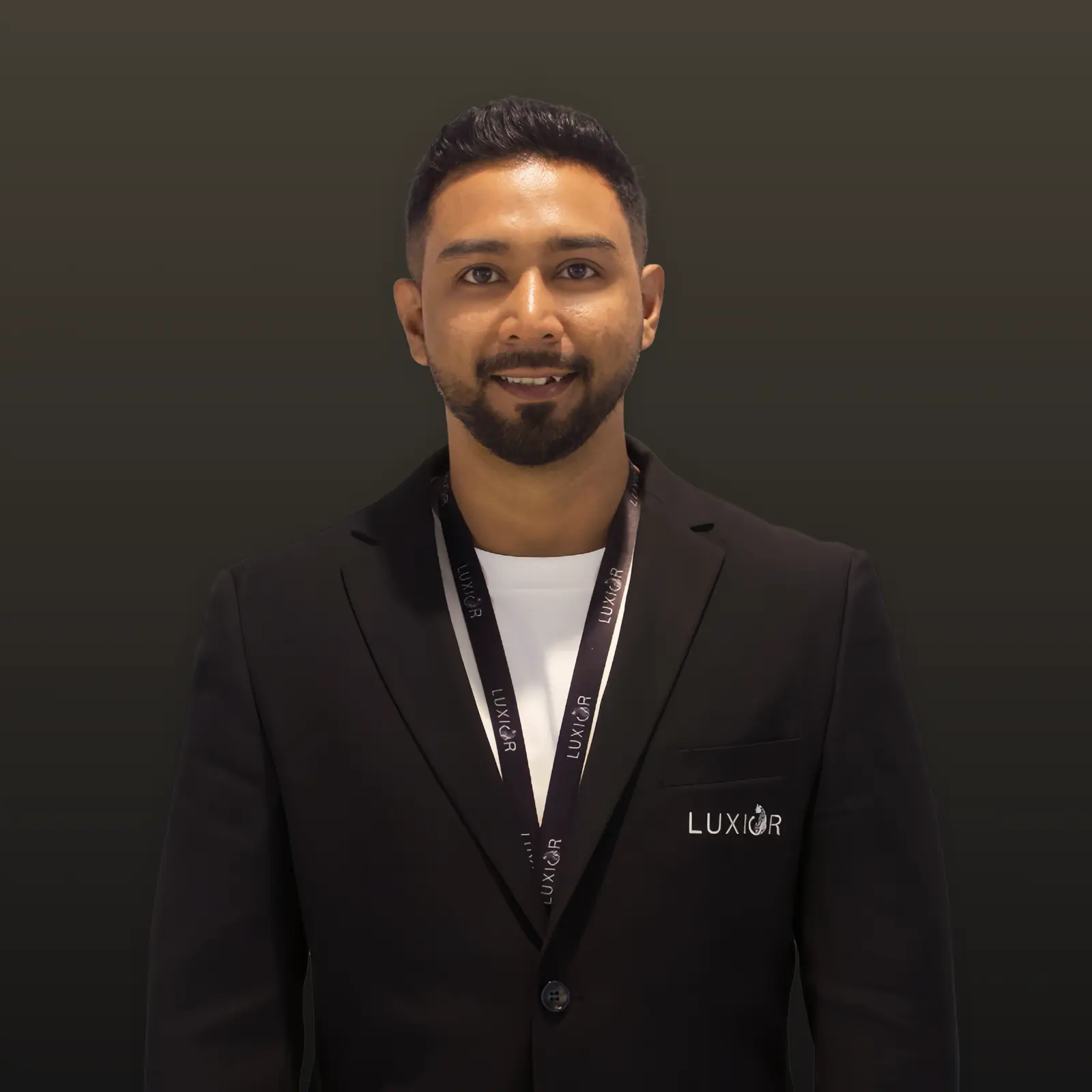 Smiling man wearing a black Luxior blazer and lanyard against a dark background.