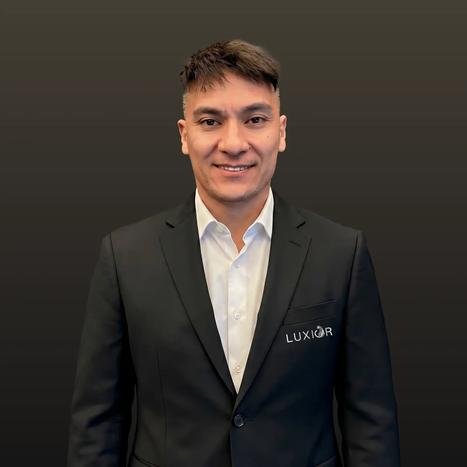 Man with short hair smiling, wearing a black LUXIOR suit jacket and white shirt against a dark background.