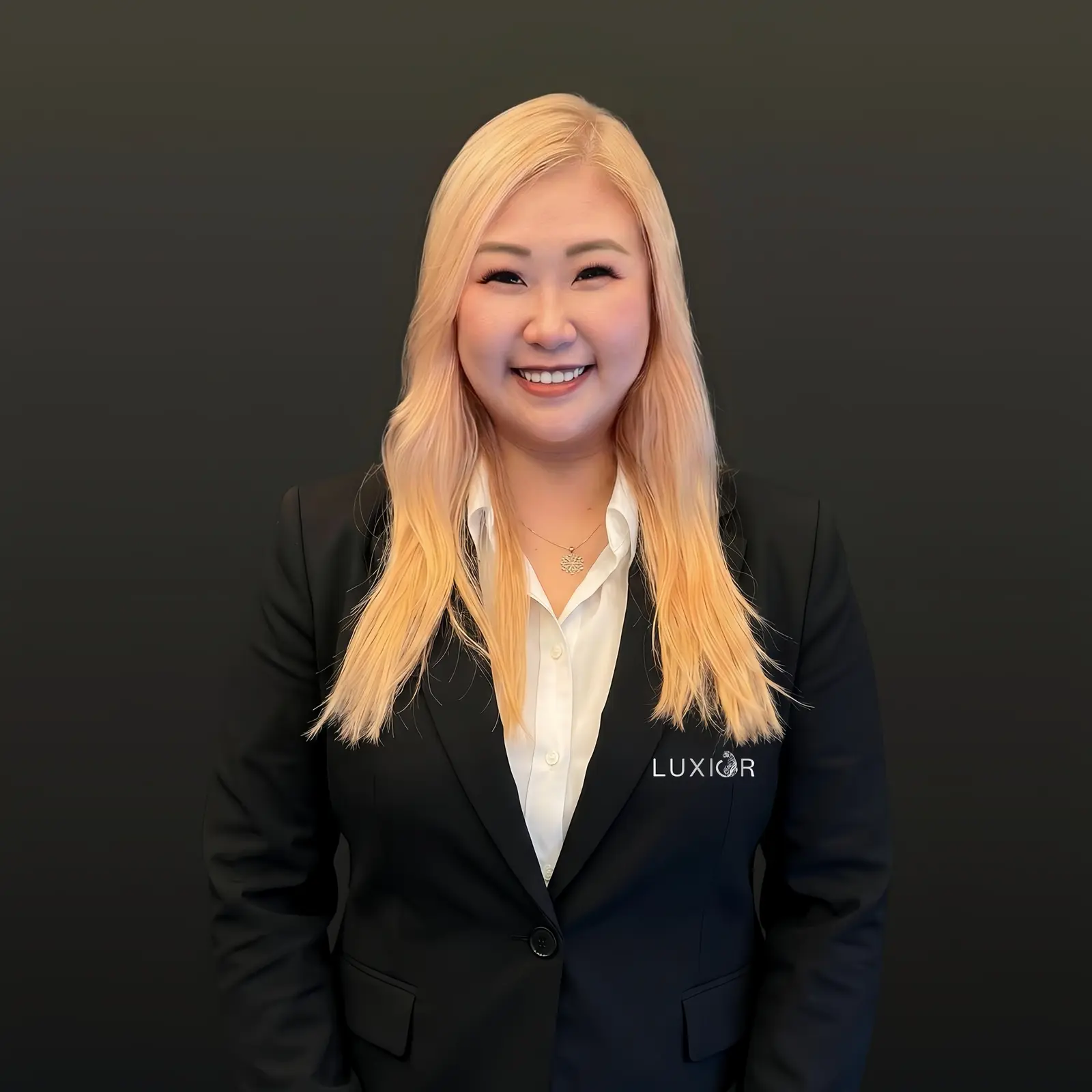 Smiling woman with long blonde hair wearing a black Luxior blazer and white shirt against black background.