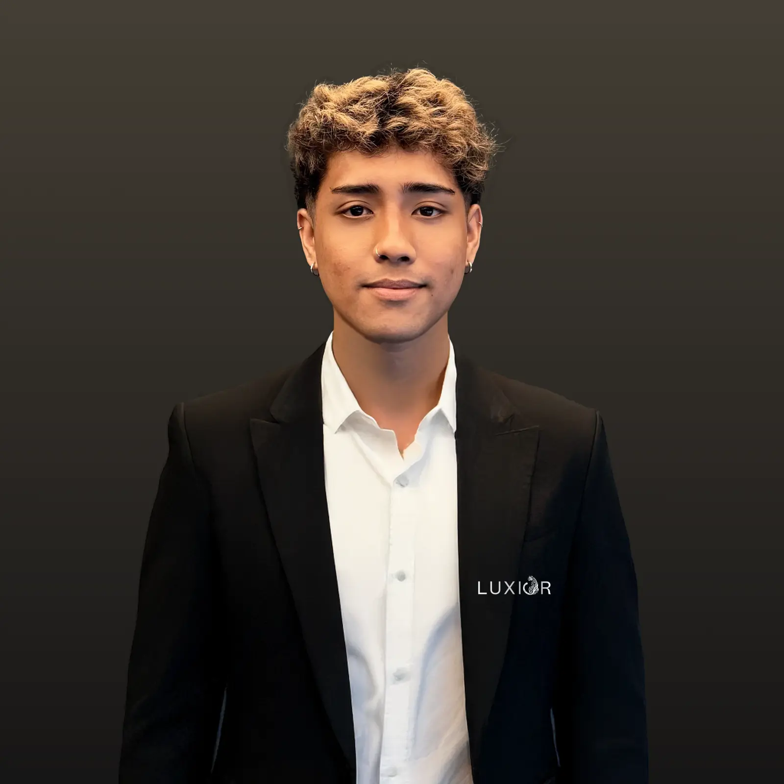Young man with curly blond hair wearing a black suit and white shirt against dark background.