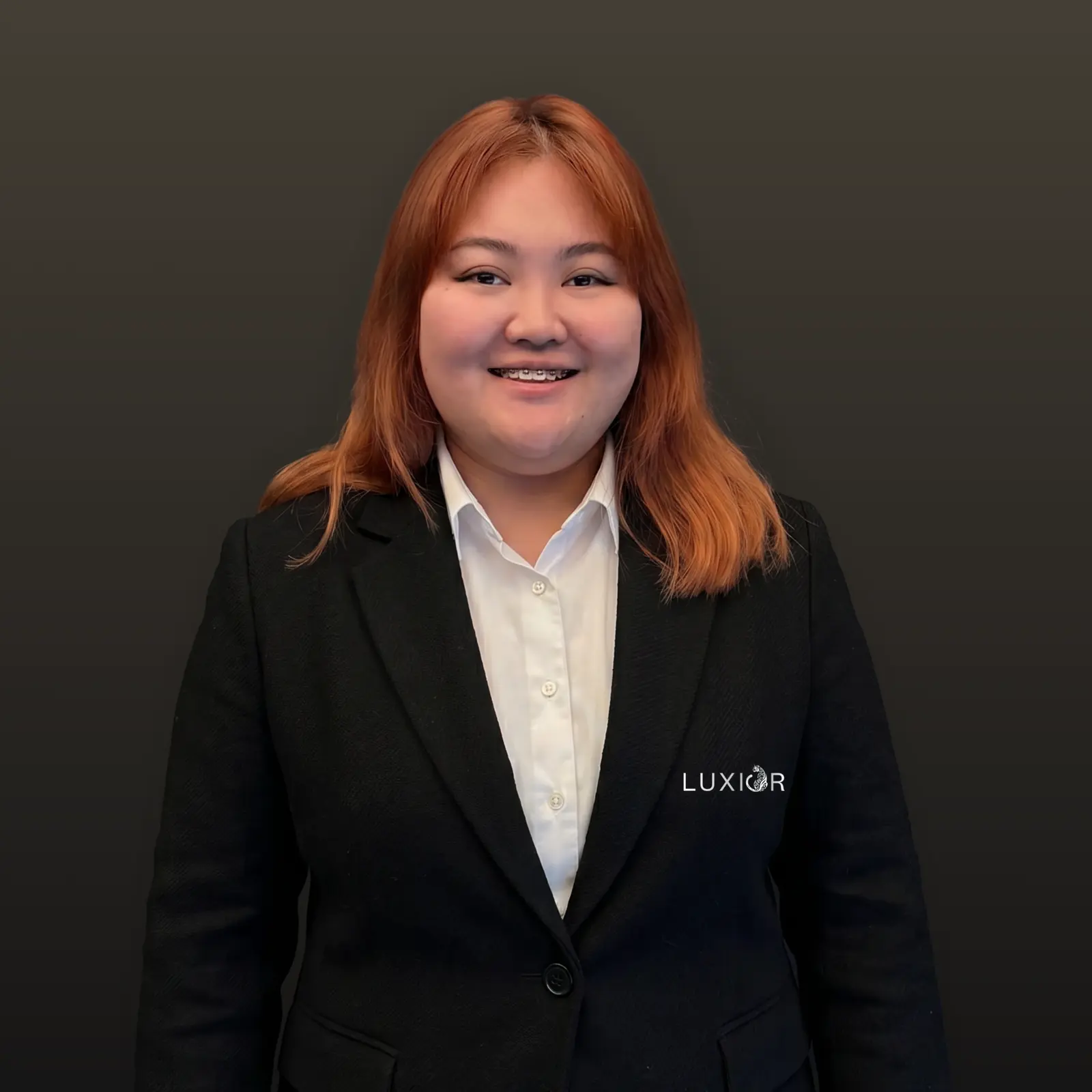 Smiling woman with reddish hair wearing a black blazer over a white shirt on a dark background.