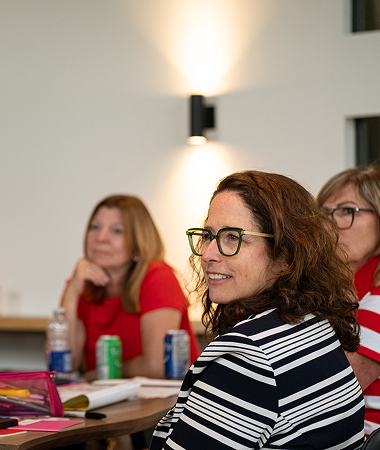 Trois femmes assises, une en avant-plan souriante portant des lunettes et un chandail rayé noir et blanc.