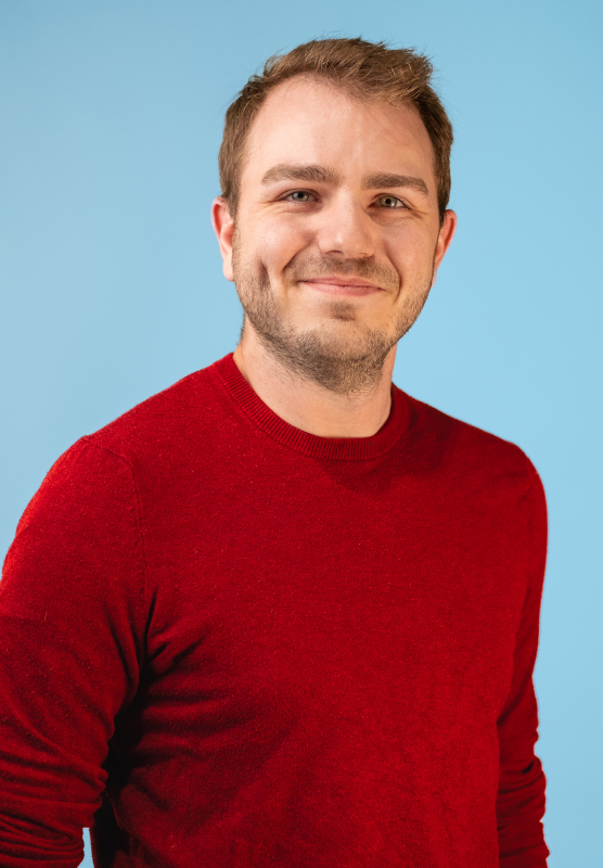 Homme souriant portant un chandail rouge brique devant un fond bleu uni.