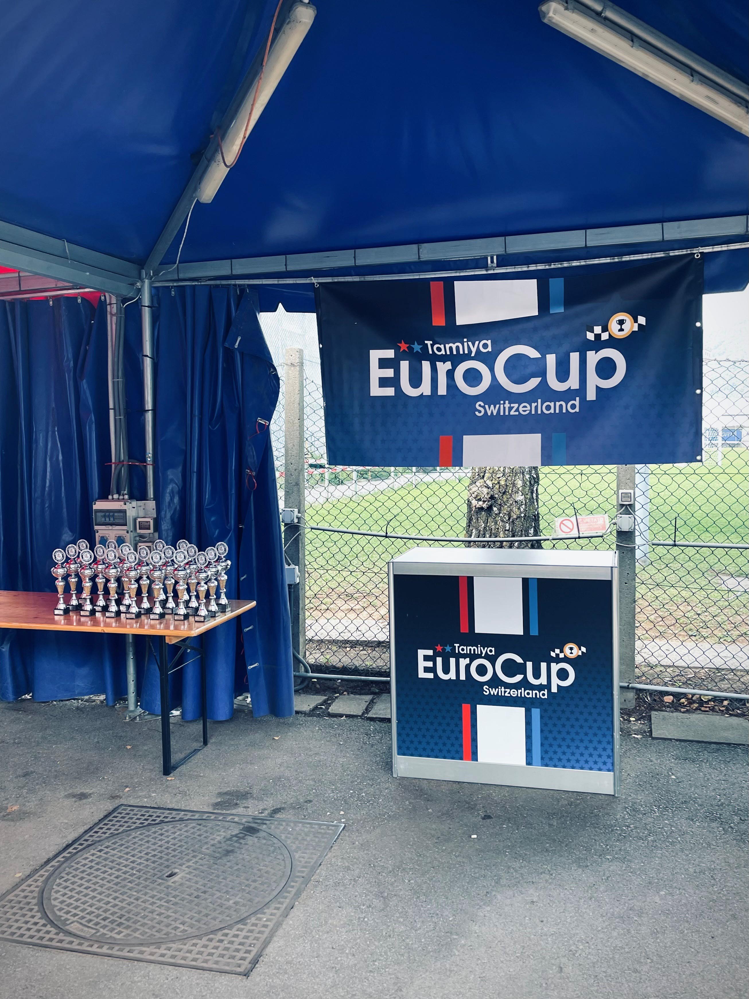 Trophies on a table under a blue tent with Tamiya EuroCup Switzerland banners displayed.