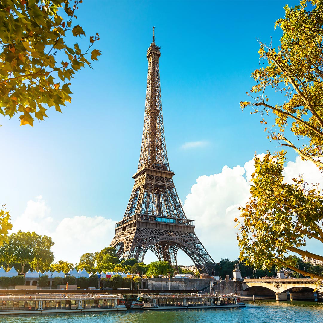Eiffel Tower in Paris with blue sky, trees, and boats on the Seine River in foreground.