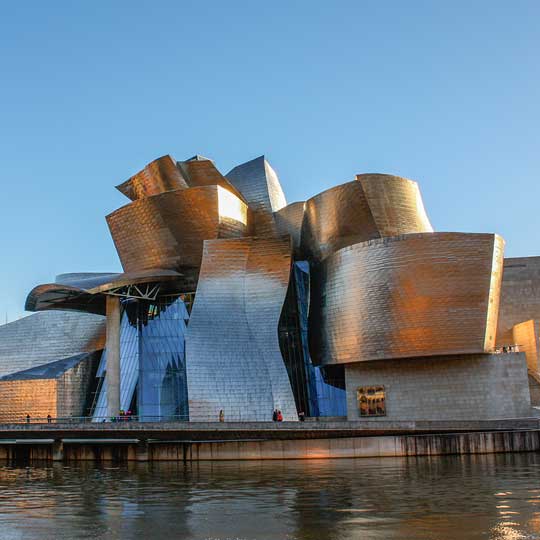 Modern metallic building with curved panels next to river reflecting its golden facade under a clear sky.