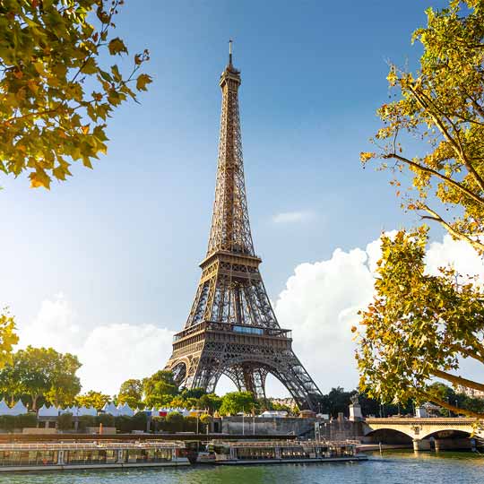 Eiffel Tower in Paris with blue sky, trees, and boats on the Seine River in foreground.