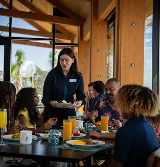 Waitress serving food to a diverse group dining at a bright restaurant with wooden beams and large windows.