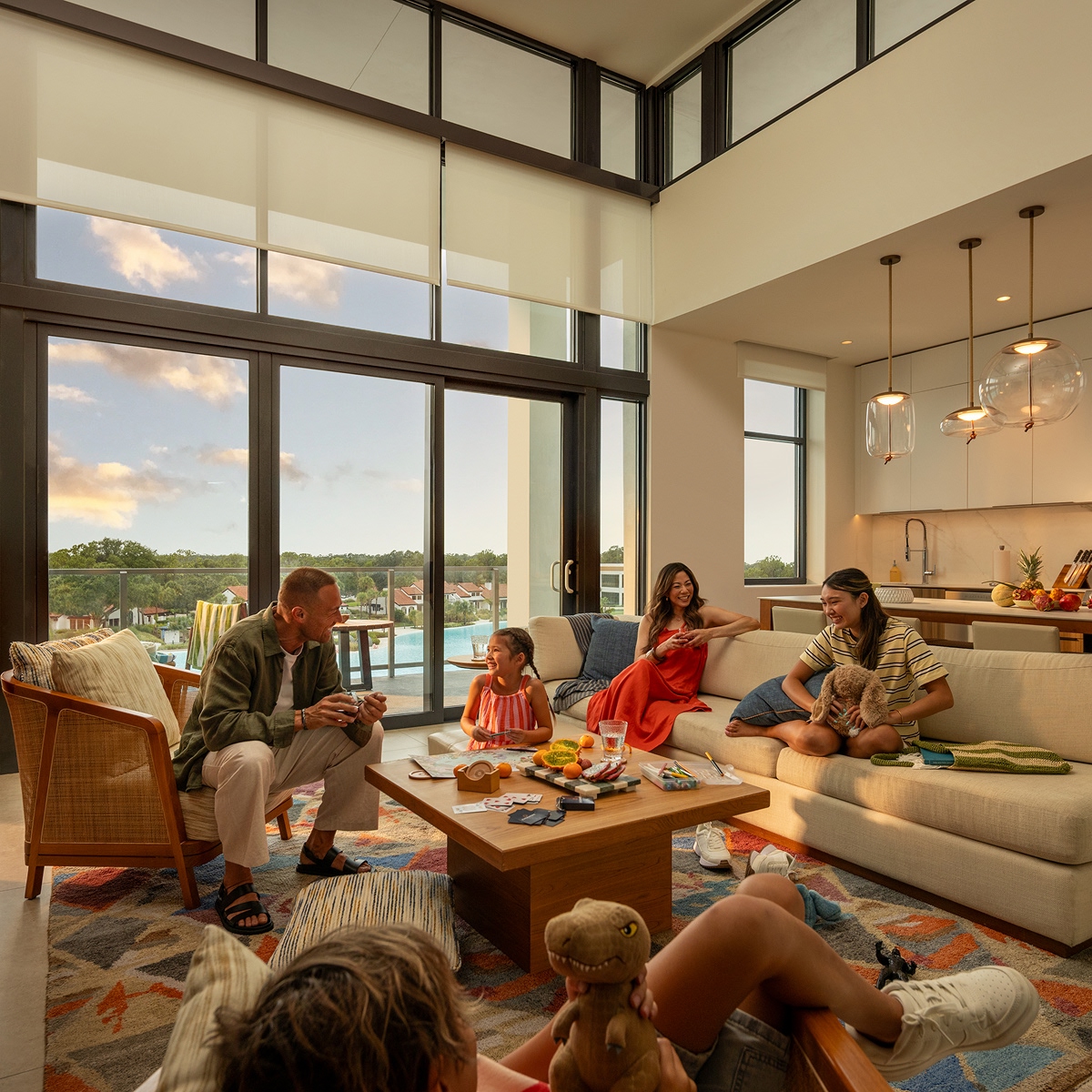 Family of five laughing and playing in a modern living room with large windows and a pool view.