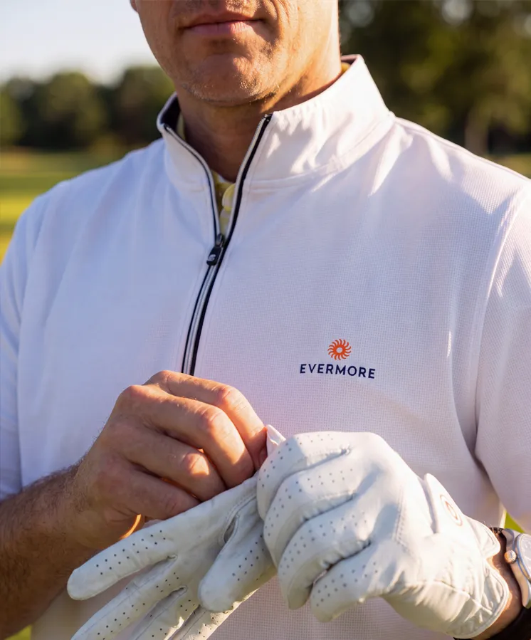Man in white Evermore polo putting on a white golf glove outdoors on a sunny day.