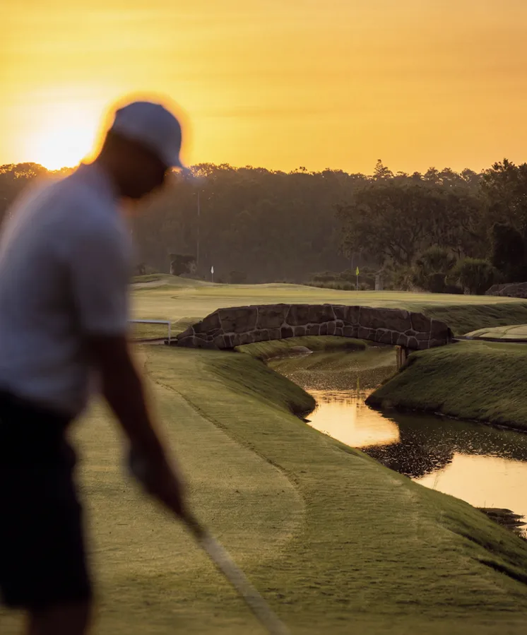 Silhouette of a golfer swinging on a golf course at sunset with a small stone bridge and water hazard.