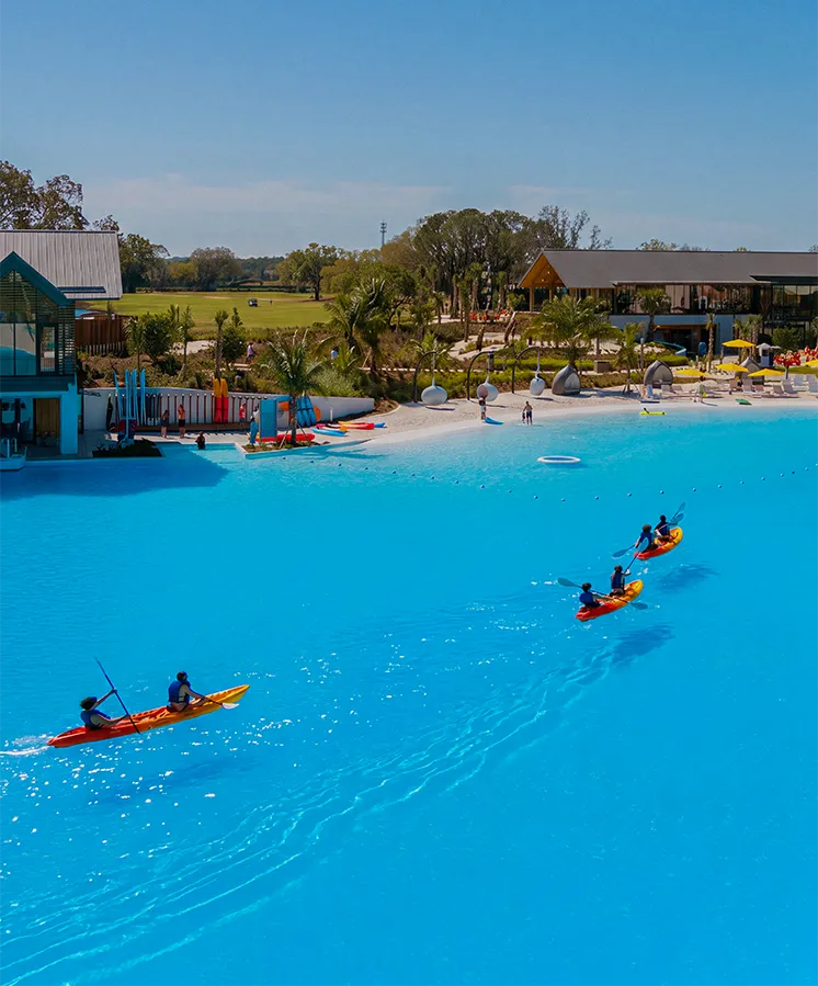Four people kayaking in a bright blue pool near a resort with palm trees and outdoor seating.