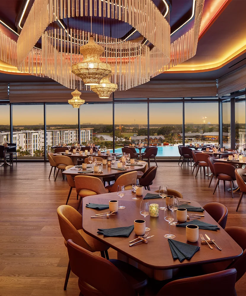 Elegant restaurant interior with wooden tables, brown chairs, large windows, and a chandelier.