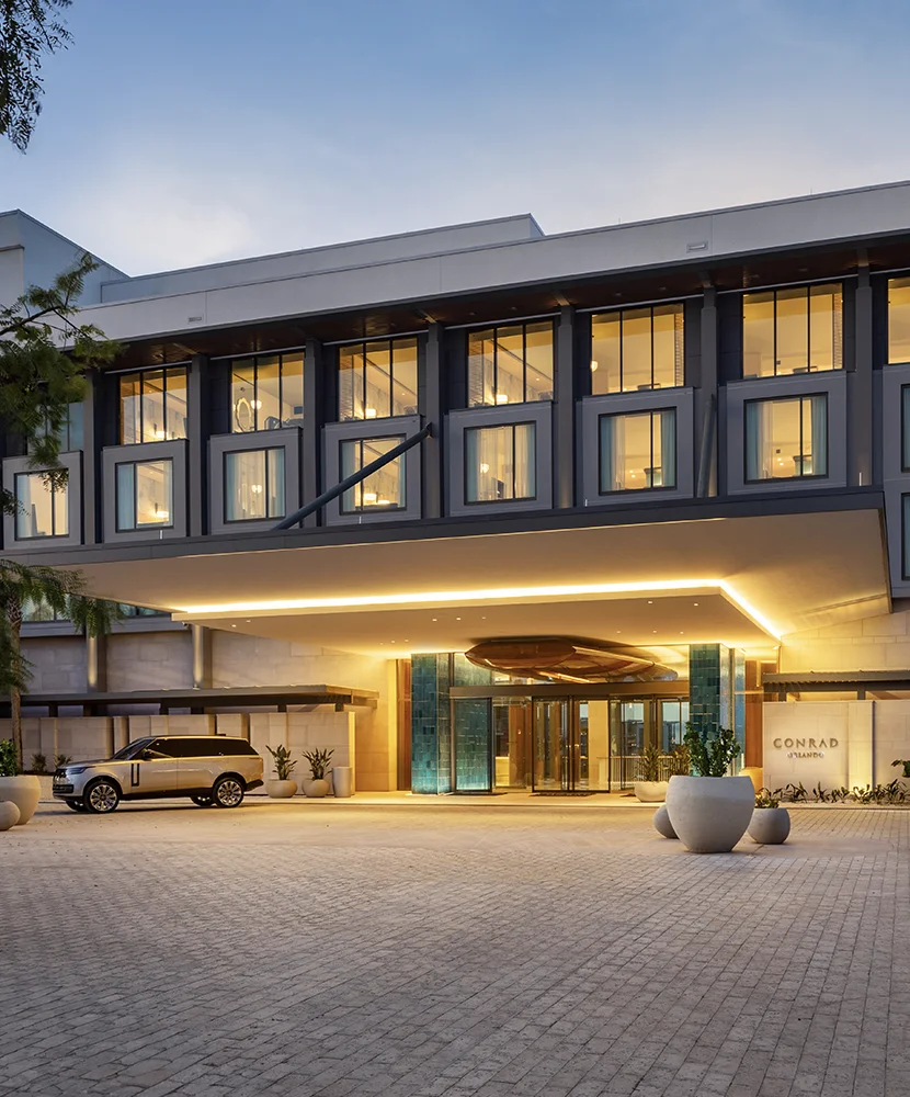 Modern hotel entrance at dusk with lit canopy, large windows, and parked silver SUV.