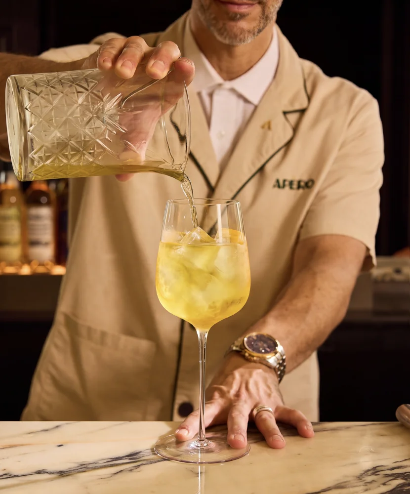 Bartender pouring a yellow cocktail with ice from a glass mixing cup into a wine glass on a marble bar.