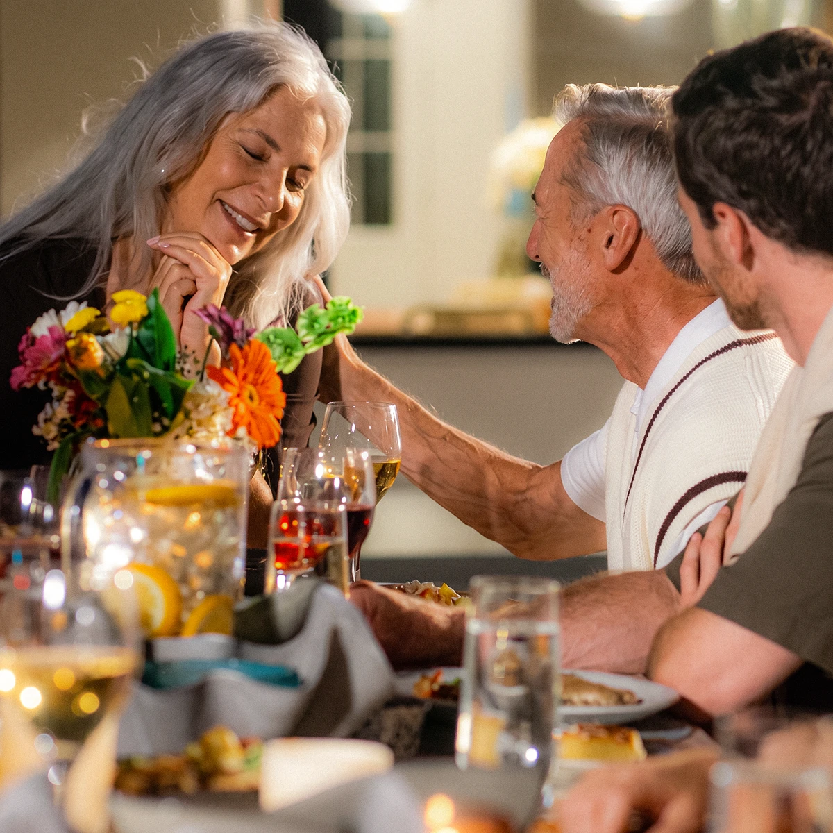 Older man gently touching smiling older woman's face at a dinner table with flowers and drinks.