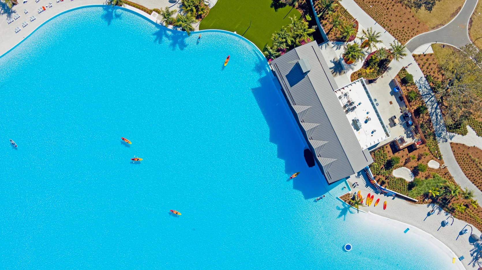 Aerial view of kayakers paddling in a large bright blue pool beside a building and landscaped area.