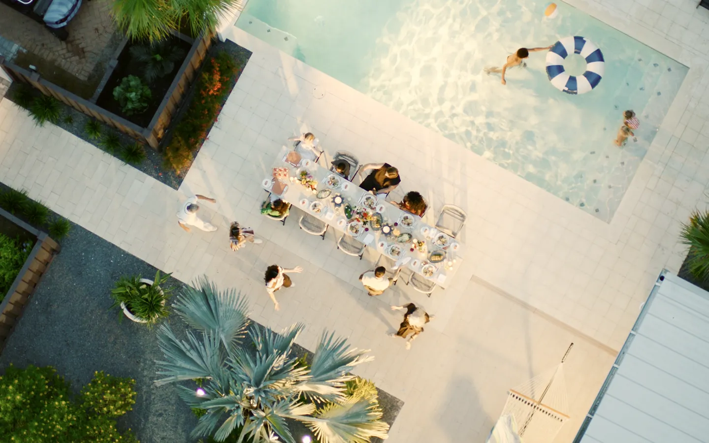 Aerial view of people dining at a table near a swimming pool with kids playing and floating ring.