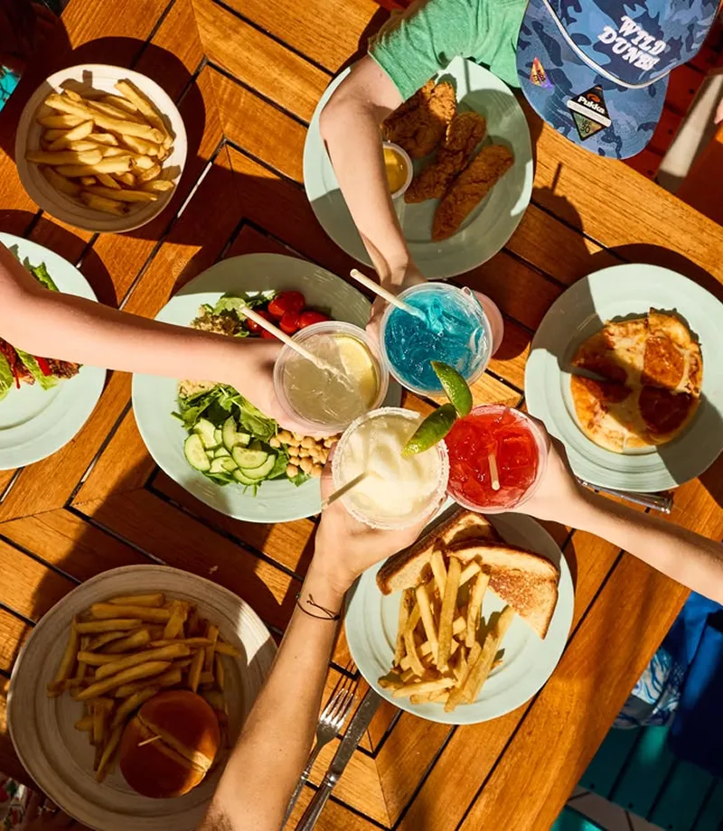 Top view of a table with plates of fries, sandwiches, salad, pizza, chicken fingers, and four hands holding colorful drinks.