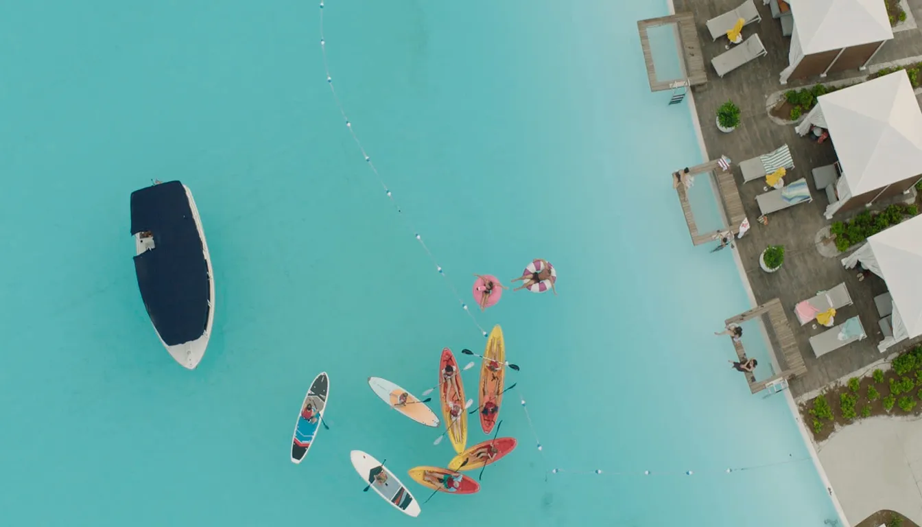 Aerial view of people paddleboarding and floating in clear blue water near a poolside with chairs and umbrellas.