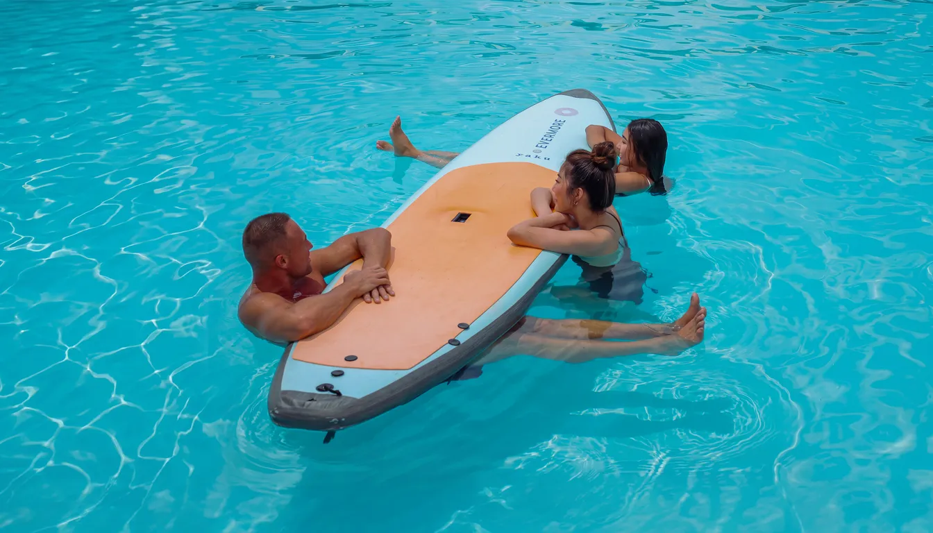 Two women and a man in a pool holding onto a large paddleboard floating on the water.
