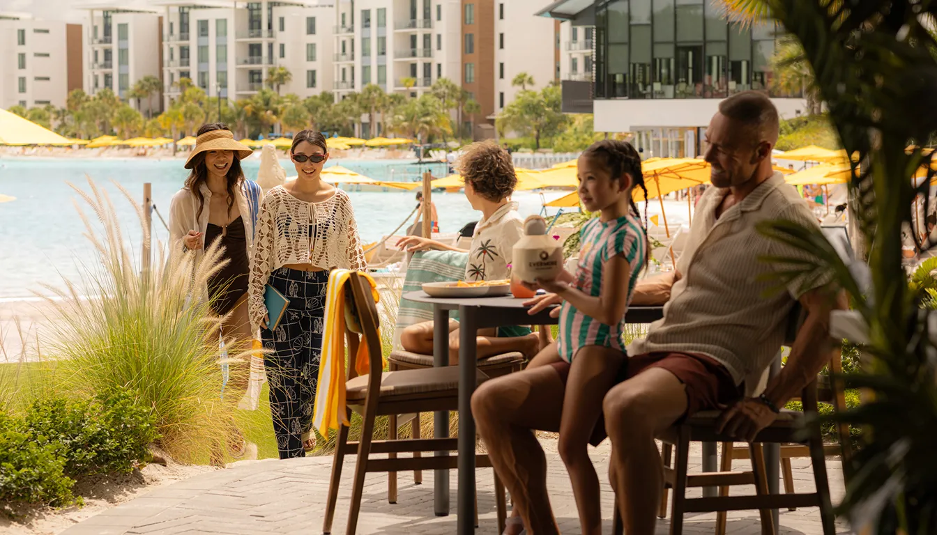 Family relaxing outdoors at a resort by the pool with guests walking nearby under yellow umbrellas.