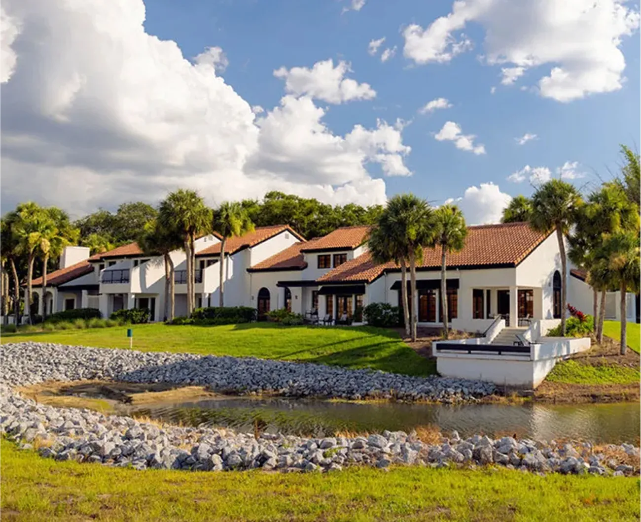 Row of white villas with red tile roofs by a small pond surrounded by green grass and palm trees.