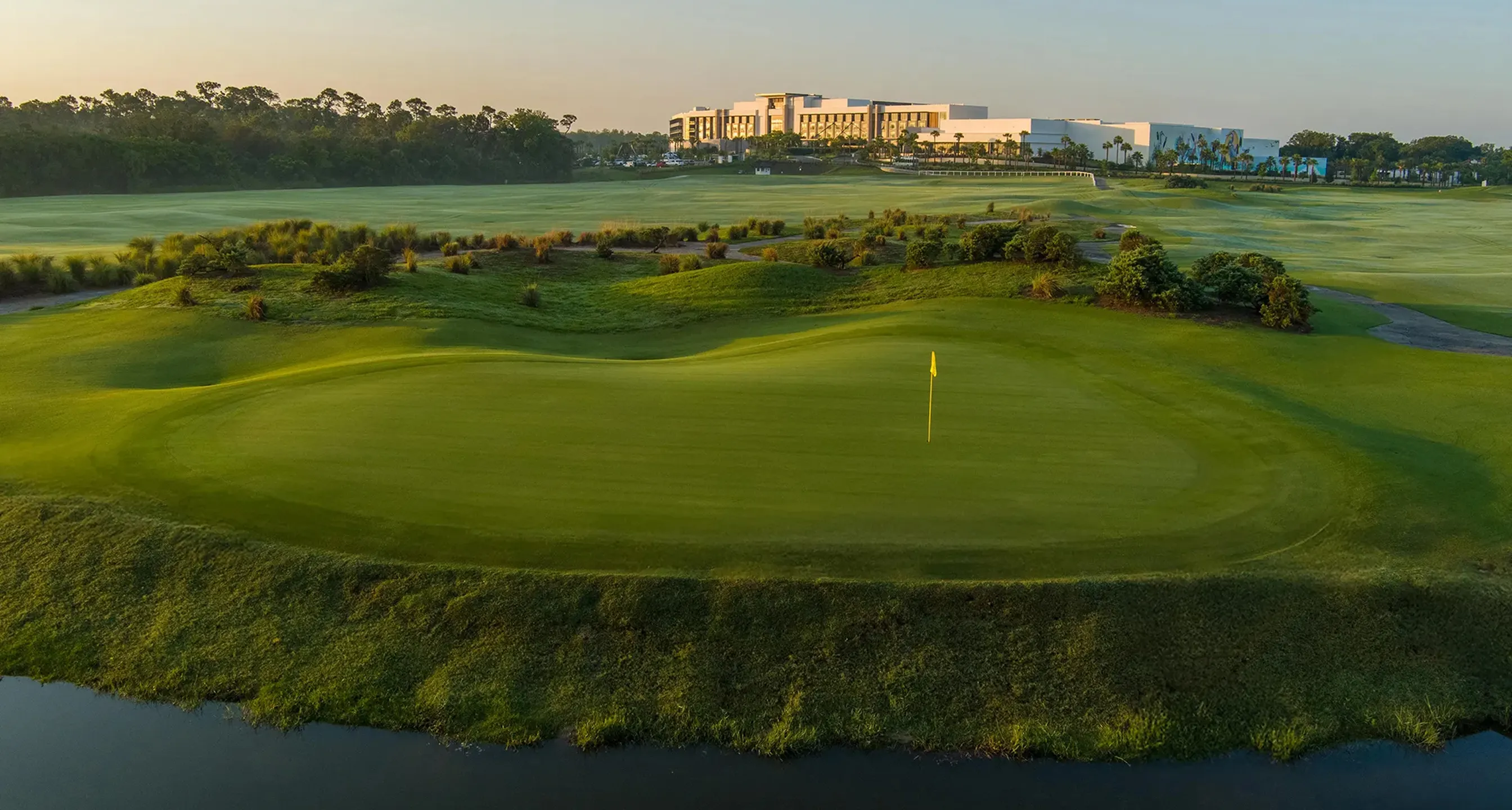 Golf green with a yellow flag, surrounded by rolling hills and water in the foreground.