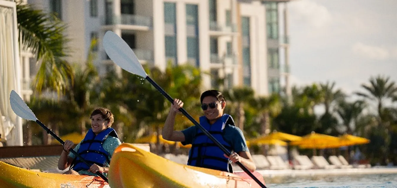 Two people kayaking in yellow kayaks near a pool with palm trees and buildings in the background.