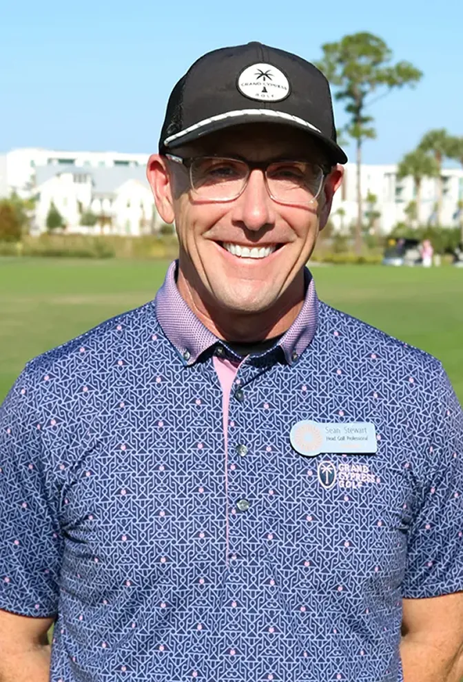 Smiling man wearing glasses, a black cap, and a patterned golf shirt on a sunny golf course.