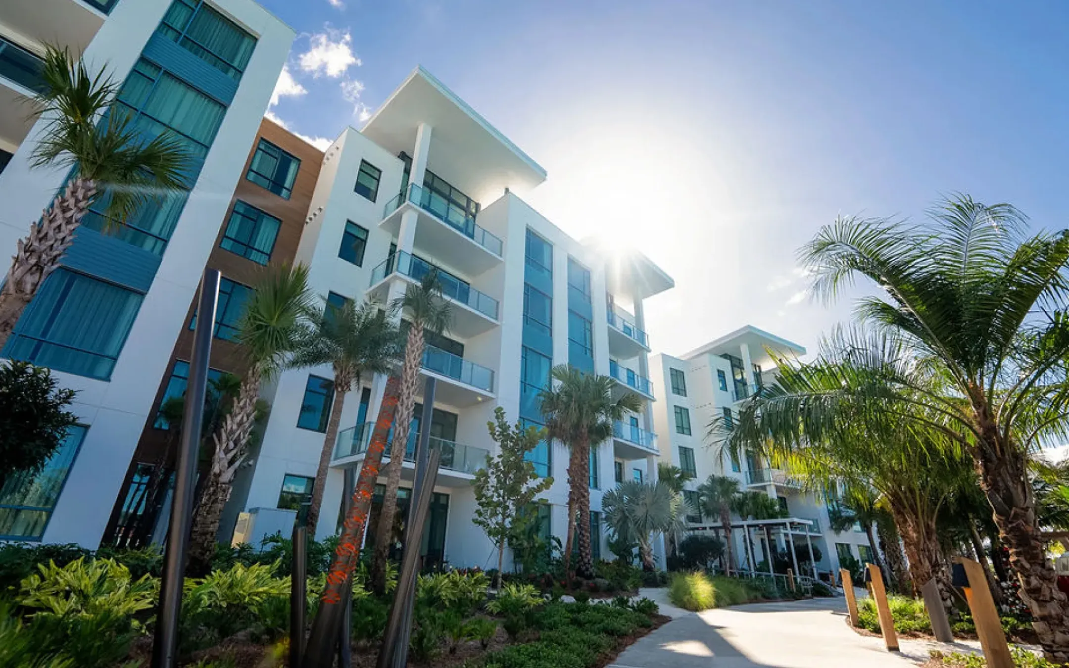 Modern apartment complex with balconies, palm trees, and landscaping under bright sunlight and blue sky.