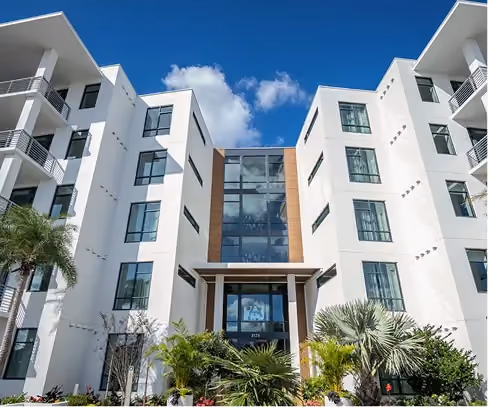 Modern white apartment building with large windows and tropical landscaping under a blue sky.