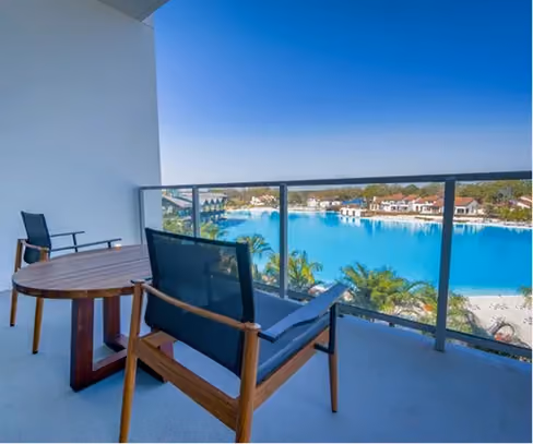 Balcony with wooden table and chairs overlooking a large blue lagoon and beachfront resort buildings.