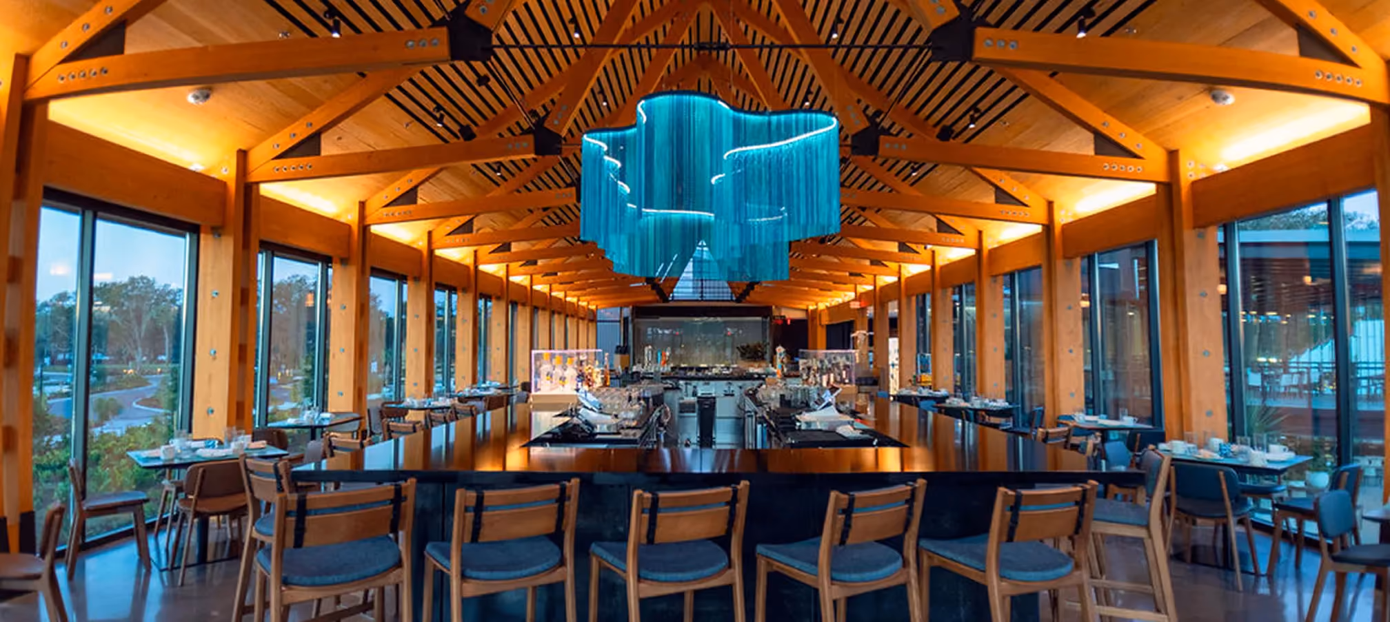 Interior of a modern restaurant with wooden beams, large windows, and a blue chandelier above the bar.