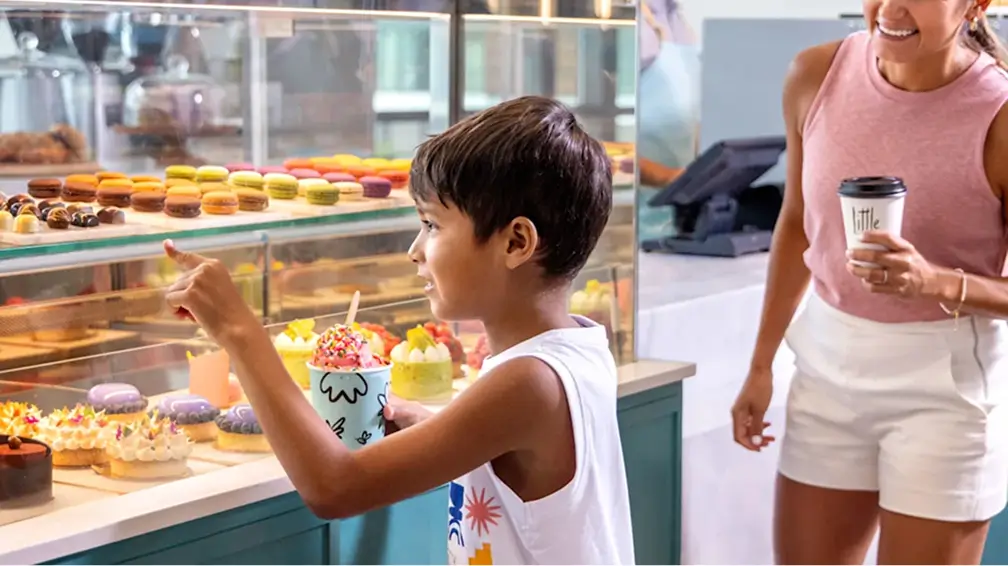 Boy holding ice cream points at macarons in a bakery display while a woman with coffee watches smiling.