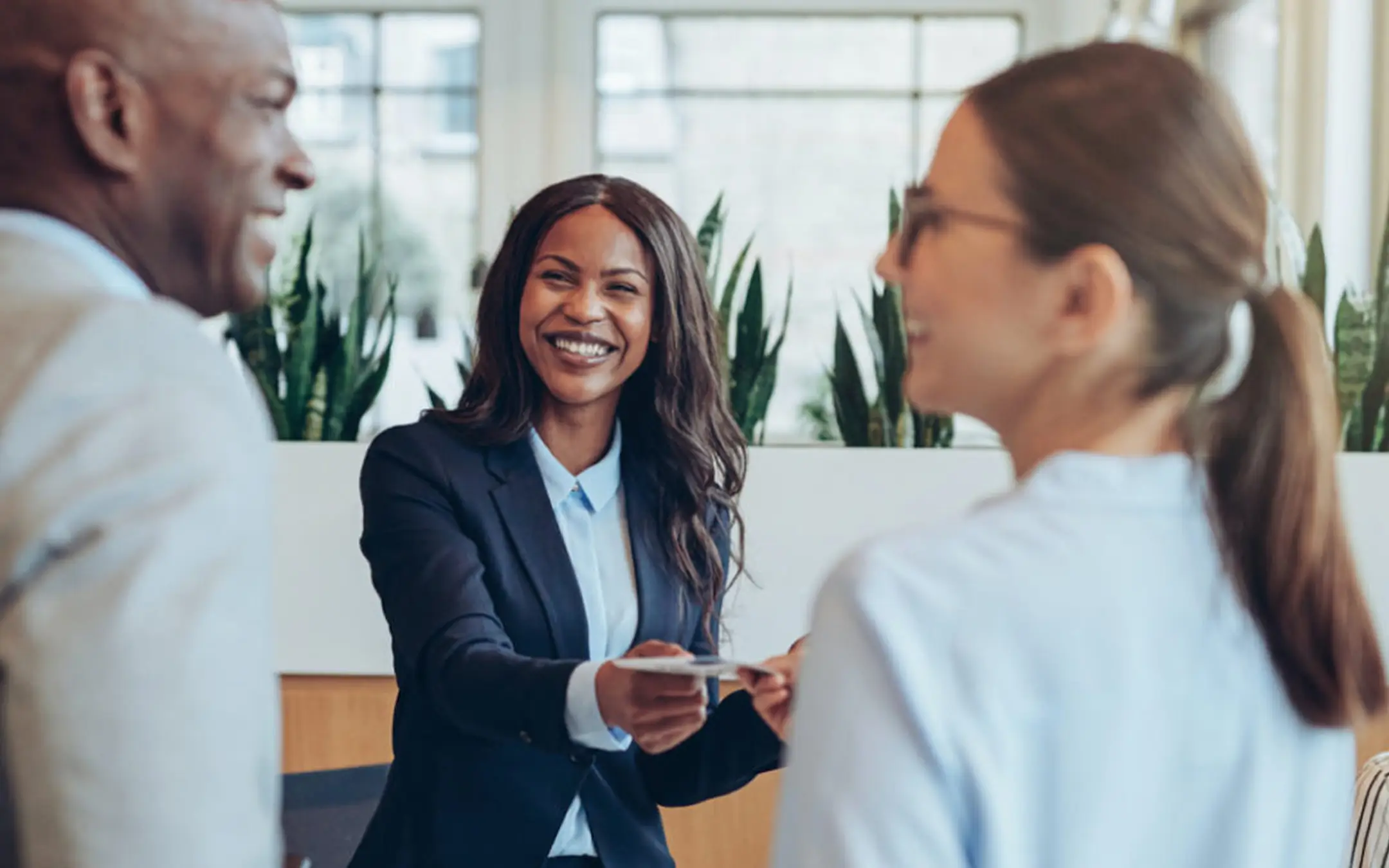 Smiling receptionist handing a card to a man and woman in a bright, modern office lobby.