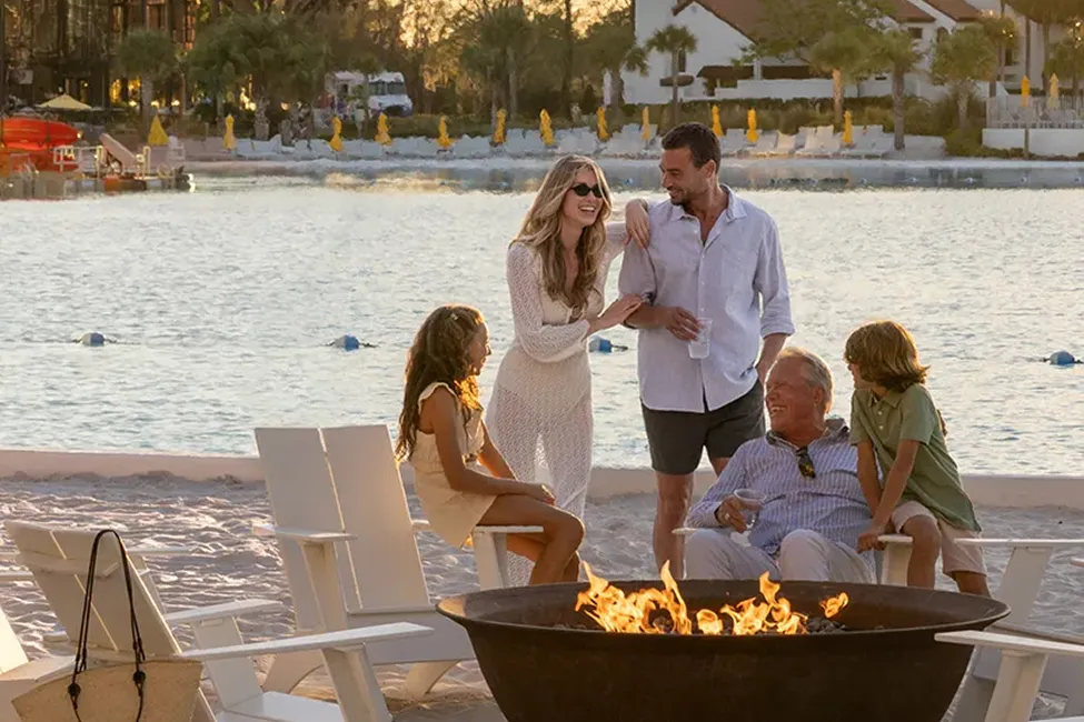 Group of people, including two kids and adults, socializing around a fire pit by a waterfront at sunset.