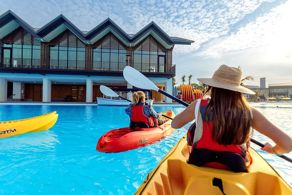 Two women kayaking on a bright blue pool near a modern building on a sunny day.