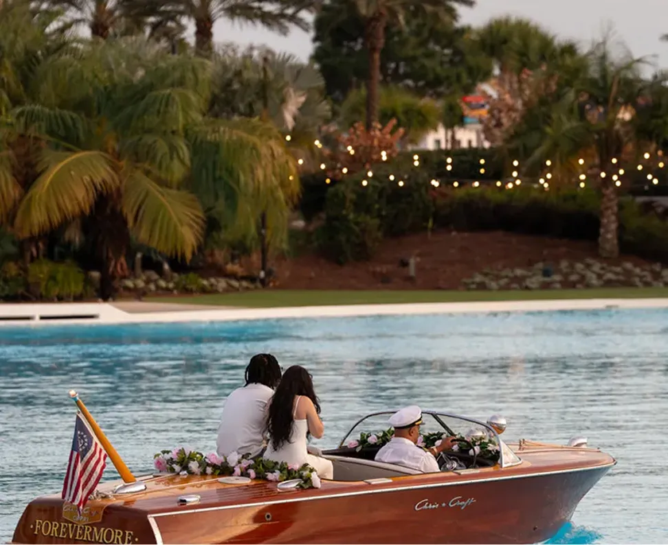 Couple sitting decorated wooden boat driven by captain on calm water with palm trees in background.