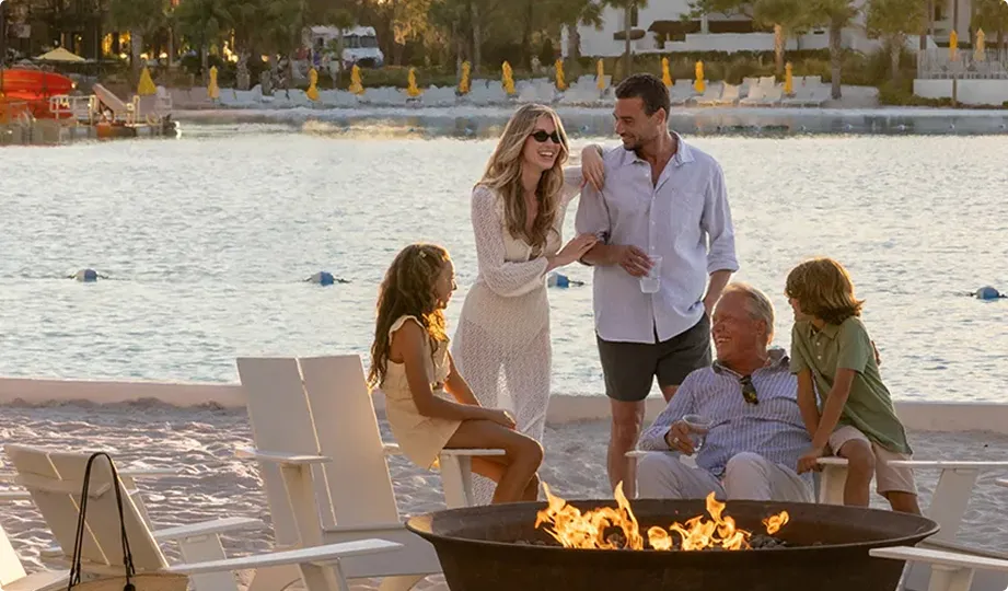 Family of five gathered around a firepit on a beach with water and lounge chairs in the background.