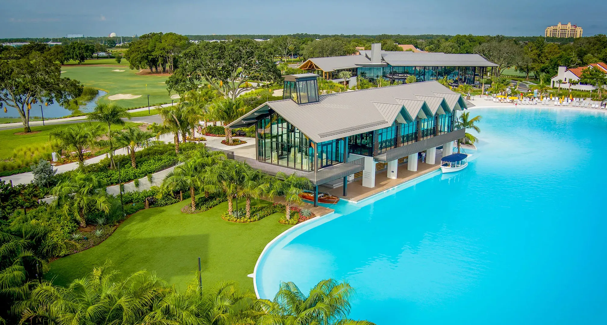 Modern glass building over a large blue pool surrounded by palm trees and greenery under a clear sky.
