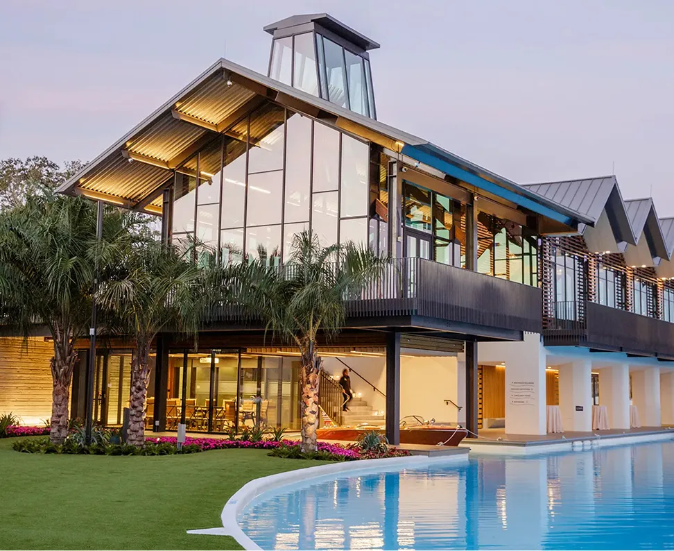 Modern glass building with palm trees beside a curved pool under a twilight sky.